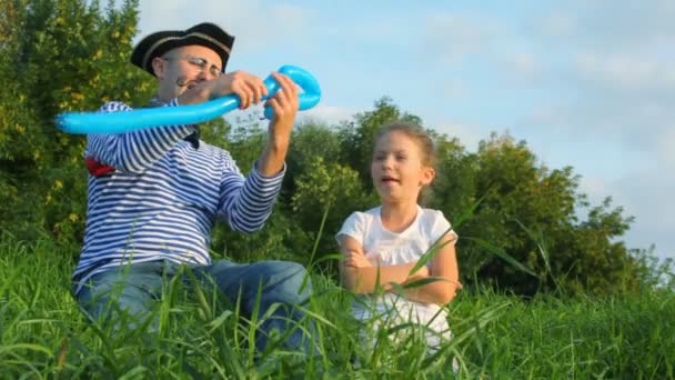 Man in piraat kostuum en meisje zit op gras en maakt zwaard uit luchtballon — Stockvideo