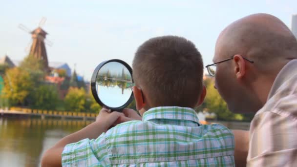 Отец и сын стоят на берегу реки и смотрят сквозь лупу — стоковое видео