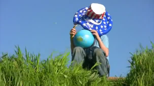 Мальчик в шляпе американского флага сидит на траве и держит земной шар — стоковое видео