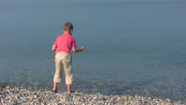 Boy stands on pebble beach and throws stones — Stock Video