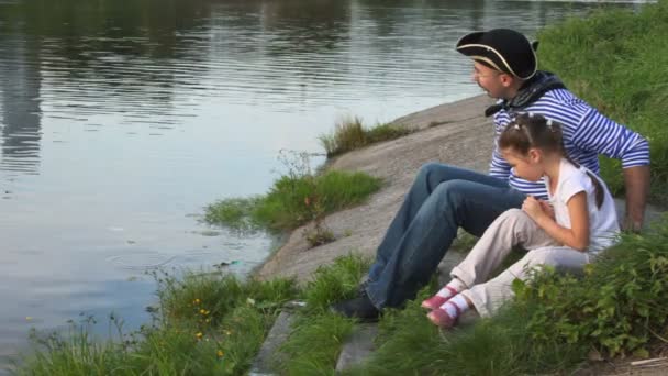 Bambina e uomo in costume da pirata si siede sulla costa e gettando ciottoli in mare — Video Stock