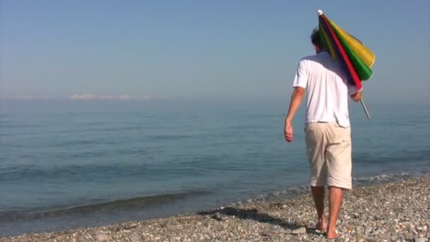 Człowiek przychodzi na plaży, dyski parasol w Kamyczki i siedzi — Wideo stockowe