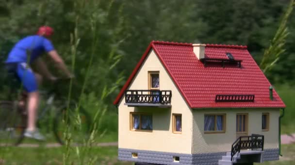 Toy house in park, unfocused group from bicyclists riding past — Stock Video