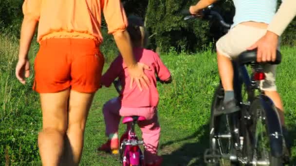 Föräldrar hjälper ett barn ridning cyklar från kameran i park — Stockvideo