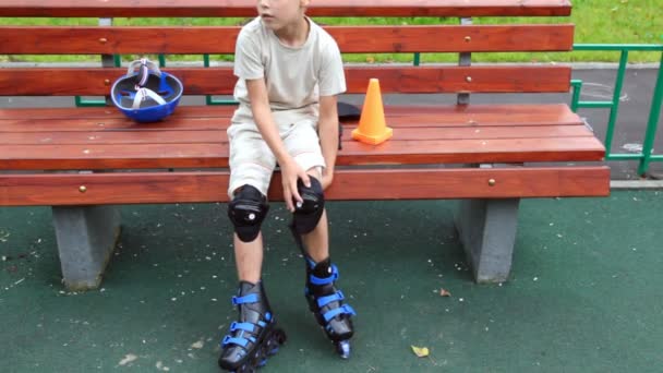 Мальчик сидит на скамейке в роликах стоя и защиты платья — стоковое видео