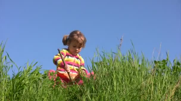 Flickan sitter på ängen och uppviglar marken och gräs med pinne — Stockvideo