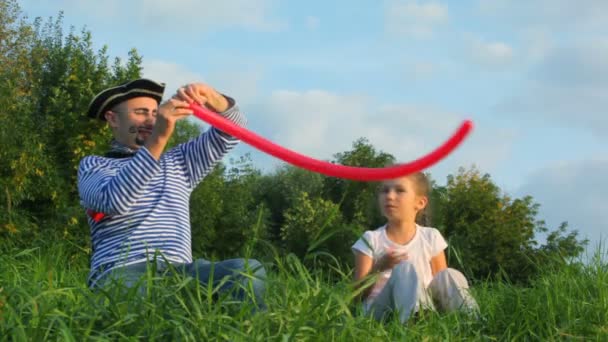 Человек в пиратском костюме дарит девушке красный воздушный шар, сидит на траве в парке — стоковое видео
