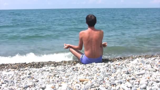Uomo seduto sulla spiaggia in posa di loto e guarda il mare — Video Stock