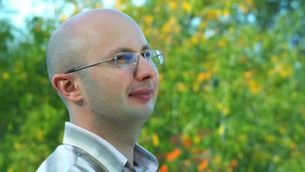 Portrait of smiling bald-headed bespectacled man stands against trees in park — Stock Video