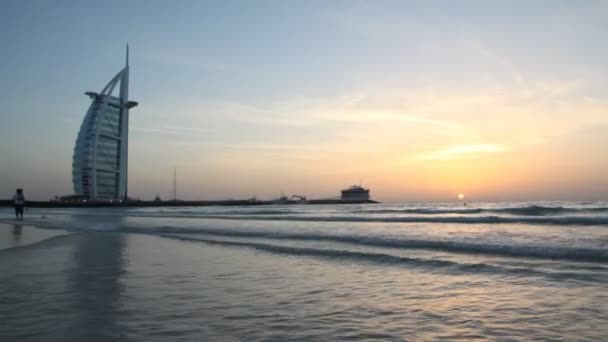 Chica corriendo en el oleaje durante la puesta del sol cerca del Burj Al Arab en Dubai — Vídeos de Stock