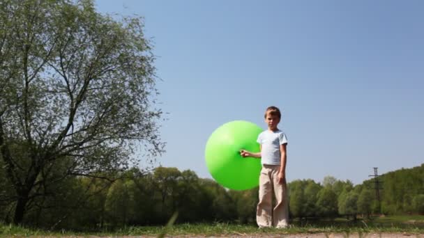 Boy holds large inflatable green ball, then carefully goes barefoot on ground and out of frame near forest in summer — Stock Video