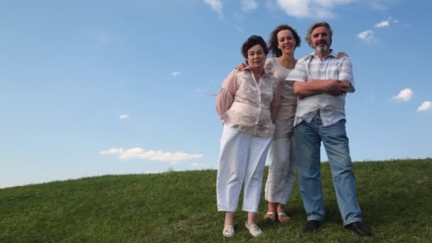 Mature couple stands embracing with their adult daughter on grass against sky in summer — Stock Video