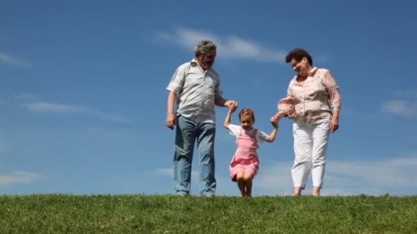 Para stoją na trawie, wnuczka skacze i kuca, trzymając się za ręce — Wideo stockowe