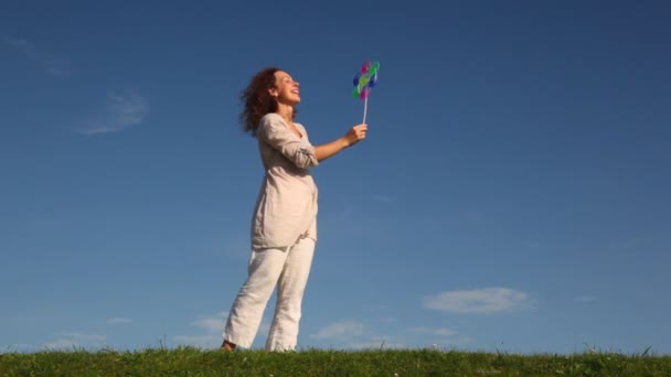 Красивая молодая женщина стоит на траве и держит в руке красочную игрушечную ветряную мельницу, вращающуюся на ветру против неба — стоковое видео
