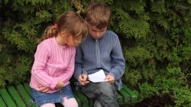 Menino e menina sentam-se no banco perto de árvores — Vídeo de Stock