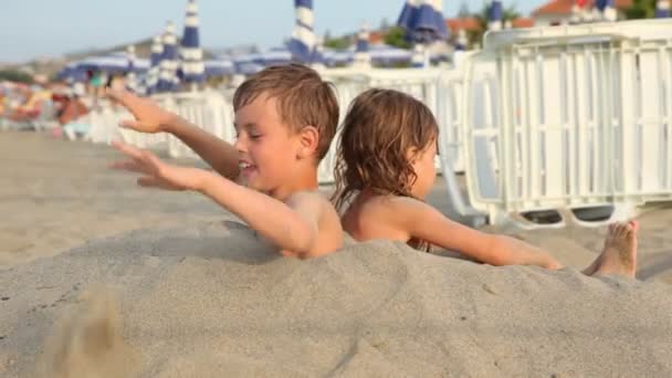 Brother and sister try to stand up buried on breast in sand on beach — Stock Video