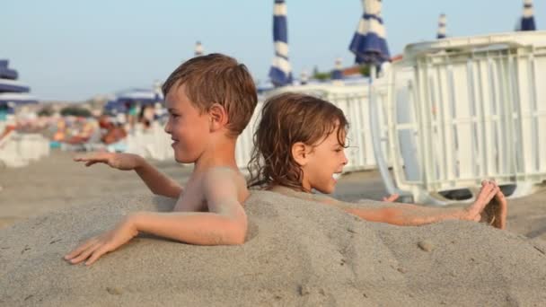 Brother and sister sit on beach buried on breast in sand — Stock Video