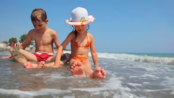 Chica y niño se sientan en el borde de la orilla del mar, jugar con la arena — Vídeo de stock