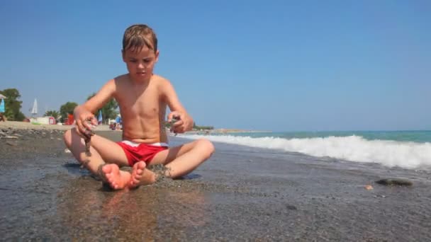 El chico se sienta al borde de la orilla del mar y juega con la arena — Vídeo de stock