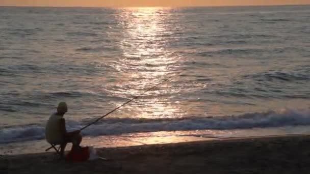 L'homme pêche et observe le coucher du soleil assis sur la côte — Video