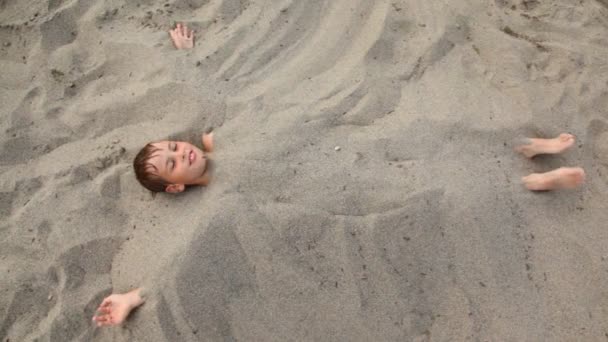 Мальчик лежит улыбается похороненный с песком на пляже — стоковое видео
