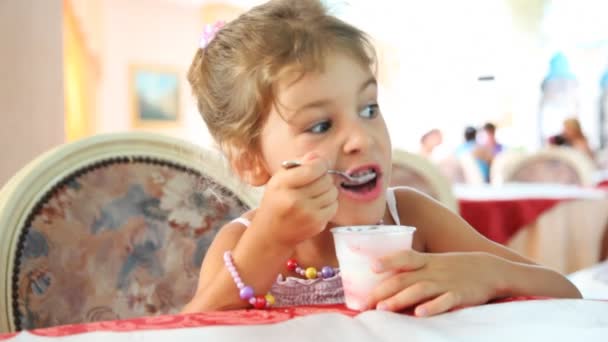 Little girl eats yogurt with spoon in plastic cup at table — Stock Video