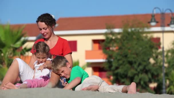 Mother sitting on sand her daughter lay between her legs and son play near — Stock Video