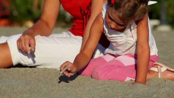 Mor och hennes dotter som sitter på sand och flickan teckning på den av pinnar — Stockvideo