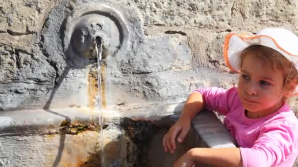 Маленька дівчинка стоїть і п'є воду зі старого джерела на стіні — стокове відео