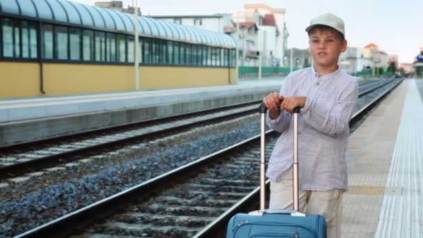 Jongen met reizen zak stands en glimlach op treinstation in de buurt van spoorlijnen — Stockvideo