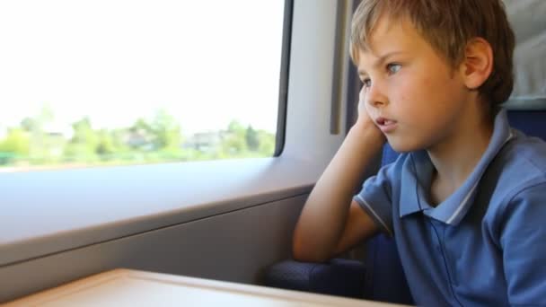 Niño se sienta y mira por la ventana de los trenes durante el movimiento se ha inclinado mejilla contra mano — Vídeos de Stock