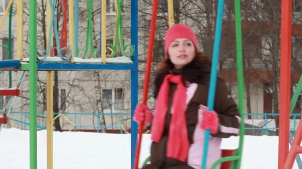 Женщина в розовой кепке и шарфе на качелях на детской площадке — стоковое видео