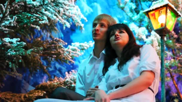 Pár sedí a dívá se nahoru poblíž barevné lucerny v zimě woods — Stock video