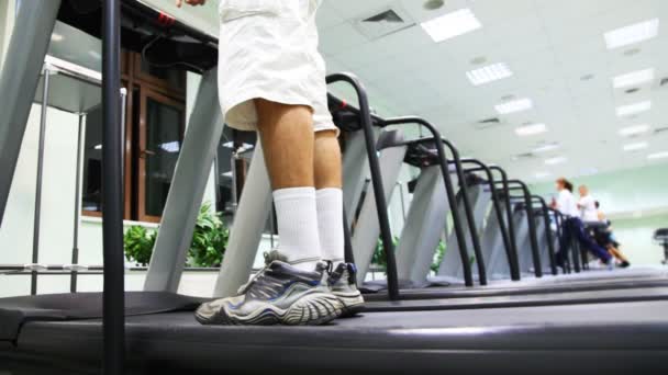 Jambes de l'homme debout au tapis roulant dans la salle de gym, il allume et commence à courir — Video