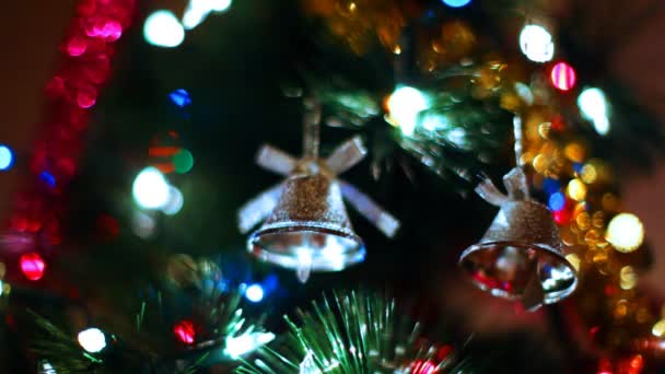 Dos campanas de juguete cuelgan en el árbol de Navidad entre guirnaldas de colores parpadeantes — Vídeos de Stock