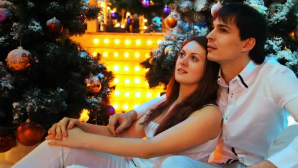 Paar zit omarmen in de buurt van besneeuwde kerstbomen en knipperende lampen — Stockvideo