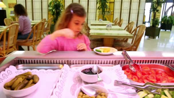 Маленькая девочка берет огурец и ставит его на тарелку в кафе — стоковое видео