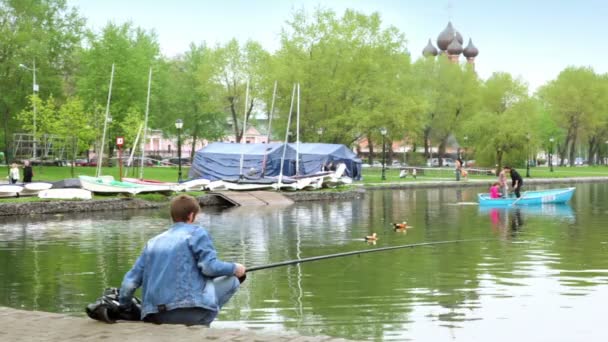 Fisherman on pond in city park, family sail on boat at background of church behind trees — Stock Video