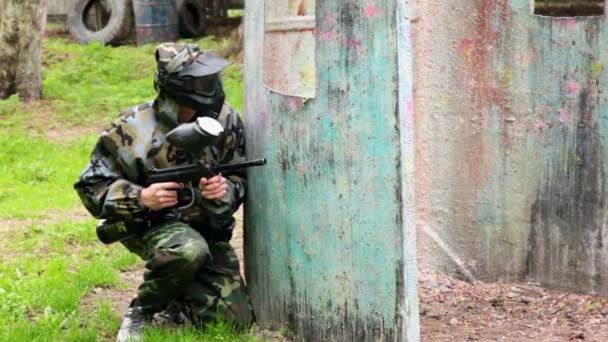 Jongen paintball speler zit in hinderlaag achter metalen hek en kijkt rond — Stockvideo