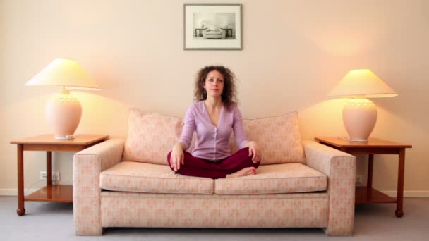 Woman sits on sofa in lotos pose, then closes her eyes and meditates — Stock Video