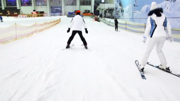 Девушка в белом платье спускается на лыжах по склону, после инструктора, который показывает упражнения — стоковое видео