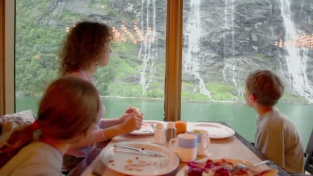 Жінка з дітьми сидить за столом і їсть перед вікном — стокове відео