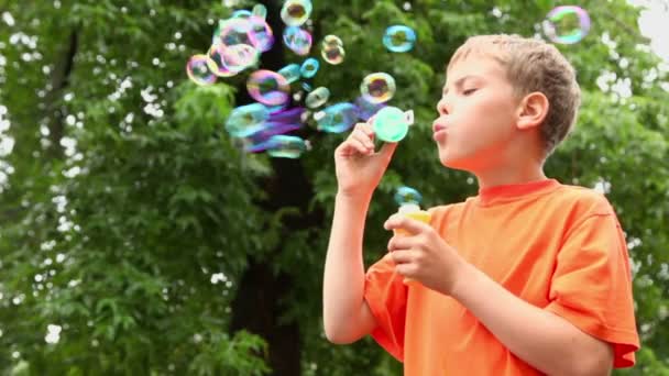 Kleine jongen staat onder regen en blaast zeepbellen in de buurt van boom — Stockvideo