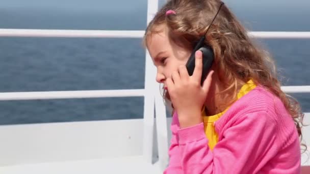 Liten flicka sitter och talar av mobiltelefon på däck nära stängslet — Stockvideo