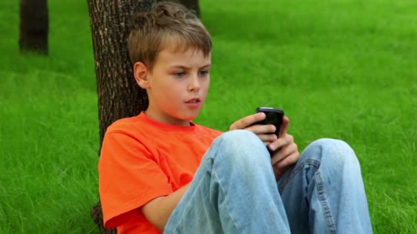 Petit garçon se penche contre l'arbre sur l'herbe et joue avec le téléphone portable 