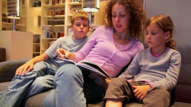 Mother with two her kids sit on sofa and read book in small room — Stock Video