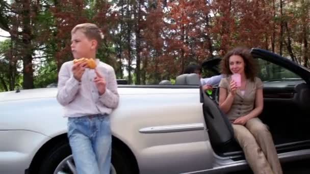 Garçon debout et mange près de cabriolet, ses parents boivent en voiture — Video