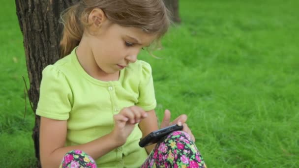 Niña se apoya en la hierba y juega con el teléfono celular — Vídeos de Stock