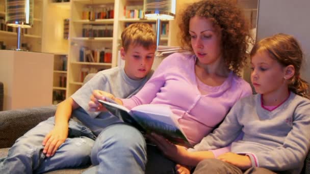 Duas crianças com a mãe sentam-se no sofá e lêem o livro no quarto pequeno — Vídeo de Stock