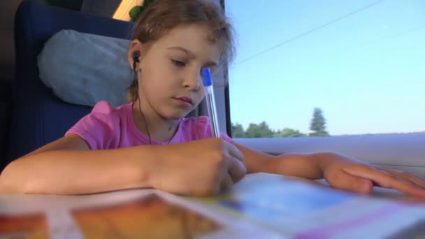 Niña dibuja por pluma en el libro cuando se sienta en el tren cerca de la ventana — Vídeos de Stock